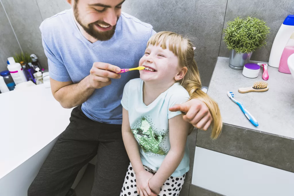 A father helping his daughter to brush teeth in a home bathroom.