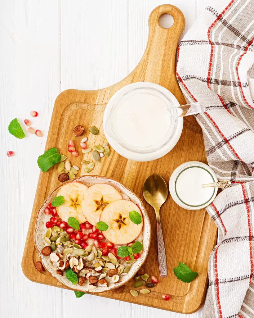 Oatmeal porridge in a bowl, decorated with apples, pomegranate, and nuts, and other utensils near it. 

