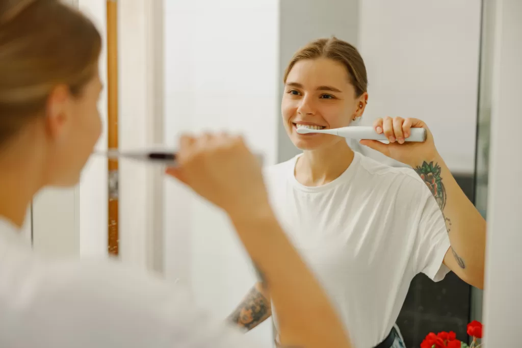 Smiling woman brushing her teeth with an electric toothbrush in front of a bathroom mirror.