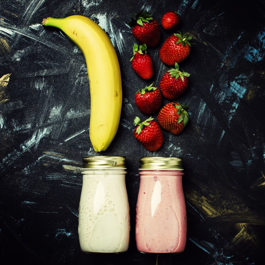 Banana and strawberry smoothies in glass jars with fresh strawberries and bananas on a dark tabletop.